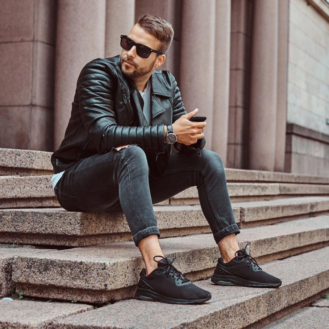 Man sitting on steps wearing sunglasses, leather jacket, and black sneakers.