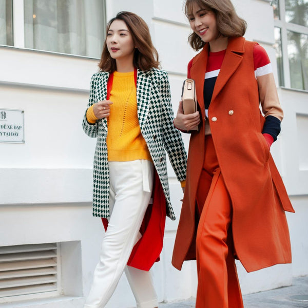 Two women walking outdoors, one in a checkered coat and yellow top, the other in an orange coat and red top.